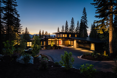 Modern house with large windows illuminated at dusk surrounded by tall pine trees and a winding driveway.