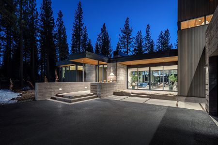 Modern house exterior with large glass windows, concrete walls, and surrounding tall pine trees at dusk.