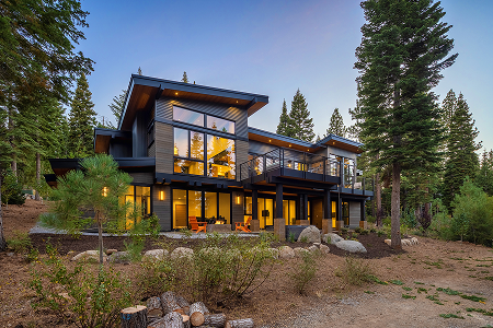 Modern two-story house with large windows and balcony surrounded by tall pine trees at dusk.