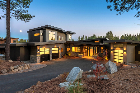Modern two-story house with illuminated windows, multiple garages, surrounded by pine trees and natural landscaping at dusk.