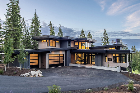 Modern two-story house with large windows and dark exterior surrounded by tall pine trees at dusk.