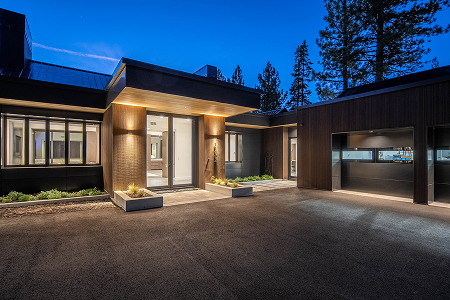 Modern house entrance illuminated at dusk with driveway and surrounding pine trees.