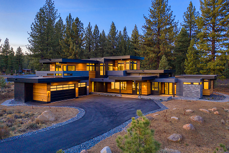 Modern two-story house with large windows and warm lighting surrounded by pine trees at dusk.