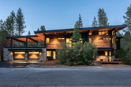 Modern two-story house with wood and stone exterior, large windows, and lit garage doors at dusk surrounded by pine trees.