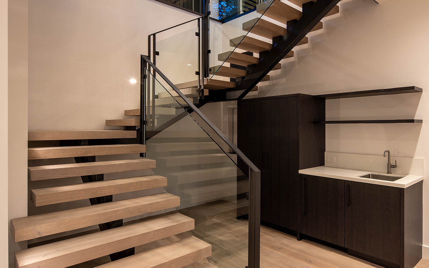 Modern interior with floating wooden stairs featuring glass railing and a dark wood kitchenette with white countertop and sink.