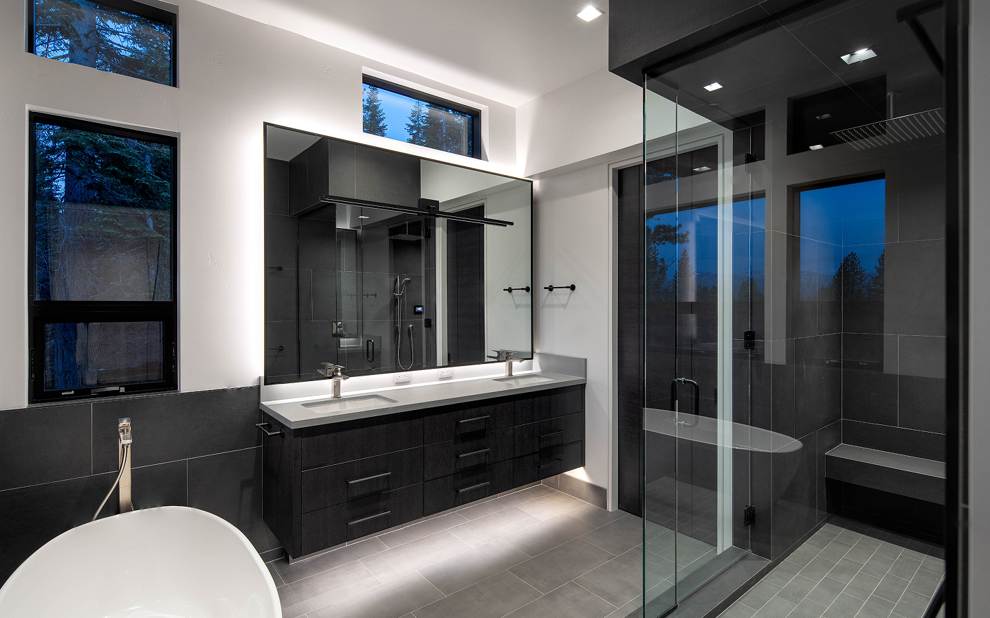 Modern bathroom with freestanding bathtub, double sink vanity with large illuminated mirror, and glass-enclosed shower with bench.