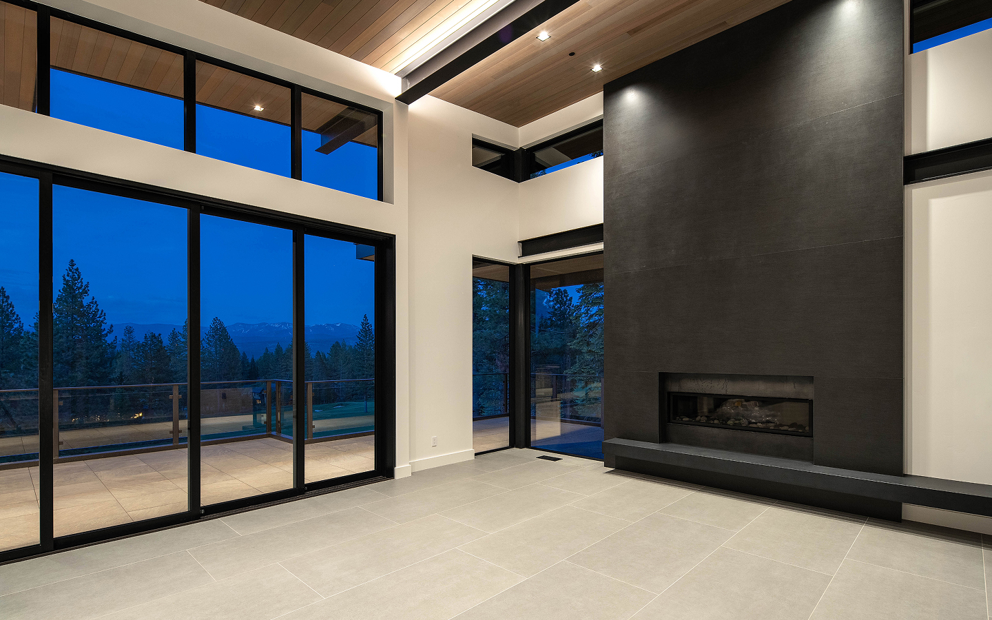 Modern living room with large glass sliding doors showing a mountain and tree view at dusk and a dark gray minimalist fireplace.