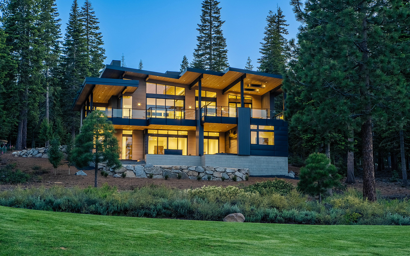 Modern two-story house with large glass windows and warm interior lighting, surrounded by tall pine trees and a green lawn.