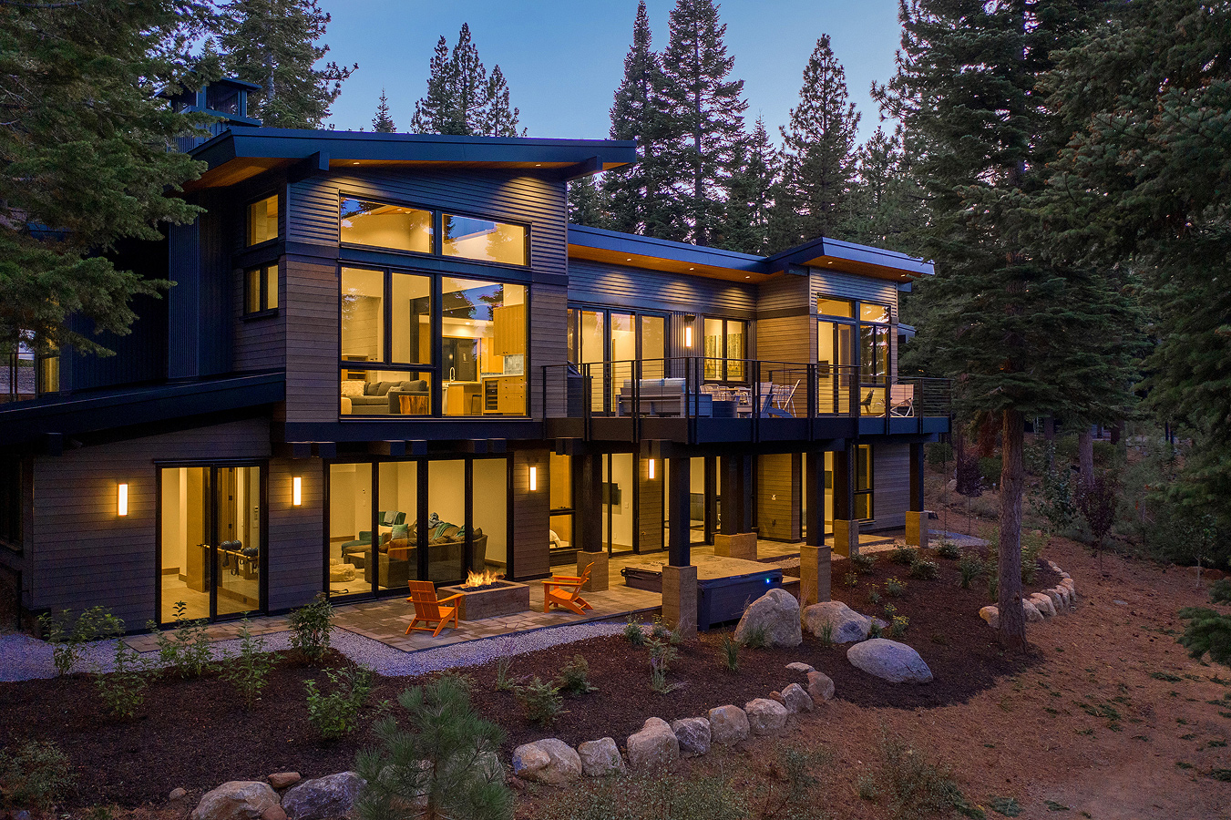 Modern two-story house with large windows illuminated at dusk, surrounded by pine trees and outdoor seating by a fire pit.