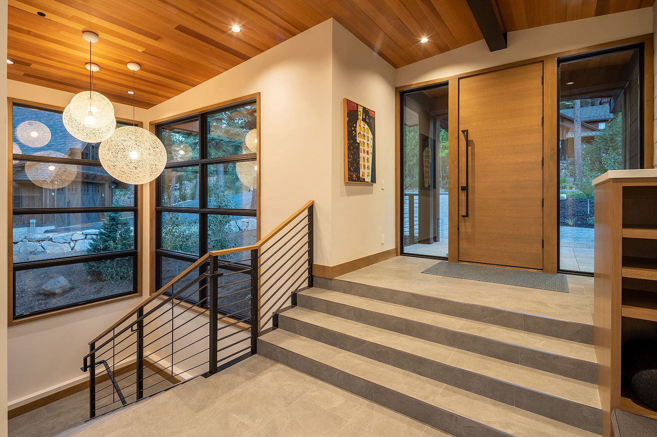 Modern home entryway with large wooden front door, tiled steps, floor-to-ceiling windows, and spherical pendant lights.