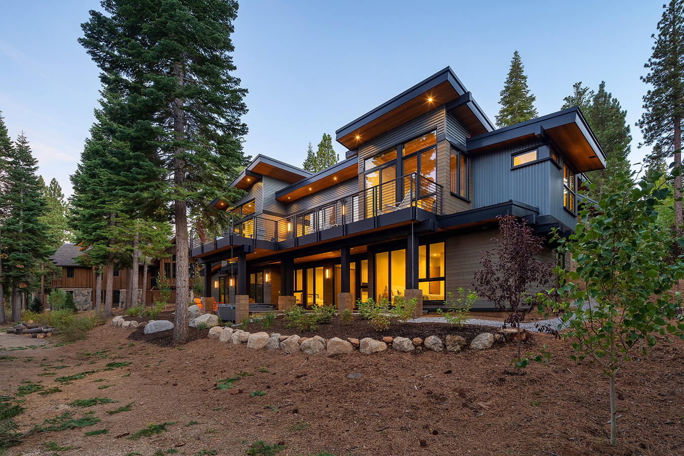 Modern two-story house with large windows lit warmly inside, surrounded by tall pine trees and landscaping with rocks and shrubs.