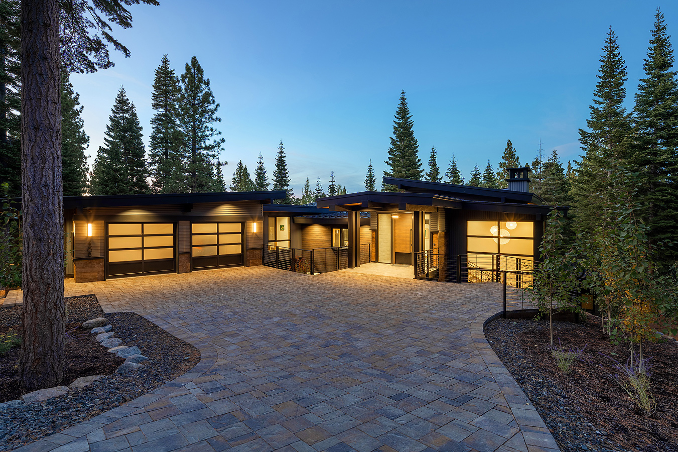 Modern single-story house with illuminated garage doors and entrance, surrounded by tall pine trees at dusk.