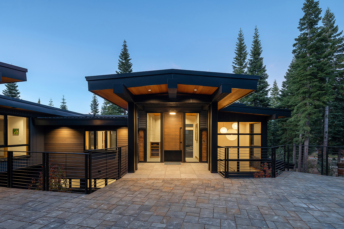 Modern house entrance with black metal frame, wood ceiling, large wooden door, and surrounding pine trees at dusk.