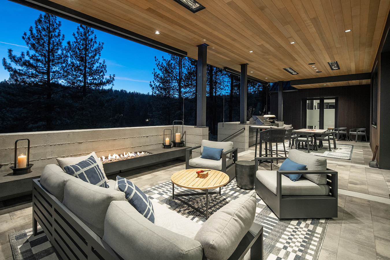 Modern outdoor patio at dusk with cushioned seating, a fire feature, wooden ceiling, and forest view.