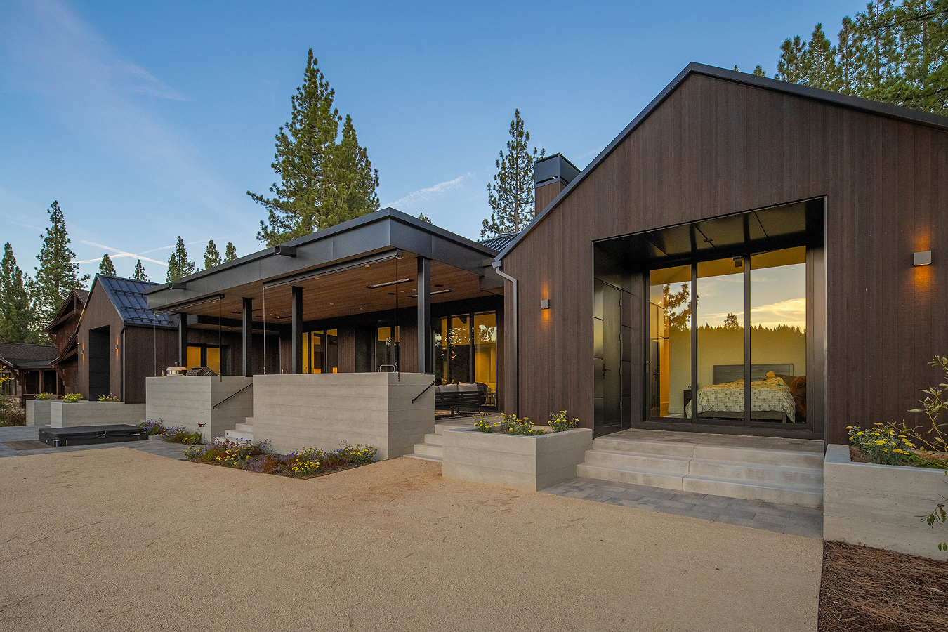 Modern wooden house with large glass windows and a concrete patio, surrounded by pine trees at sunset.