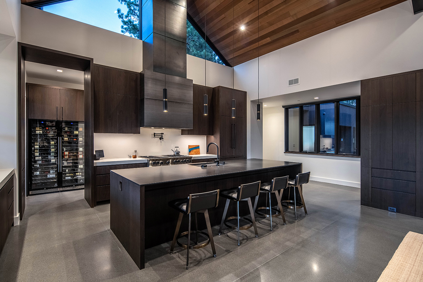 Modern kitchen with dark wood cabinets, large island with black countertop and four bar stools, high wooden ceiling, and large windows.