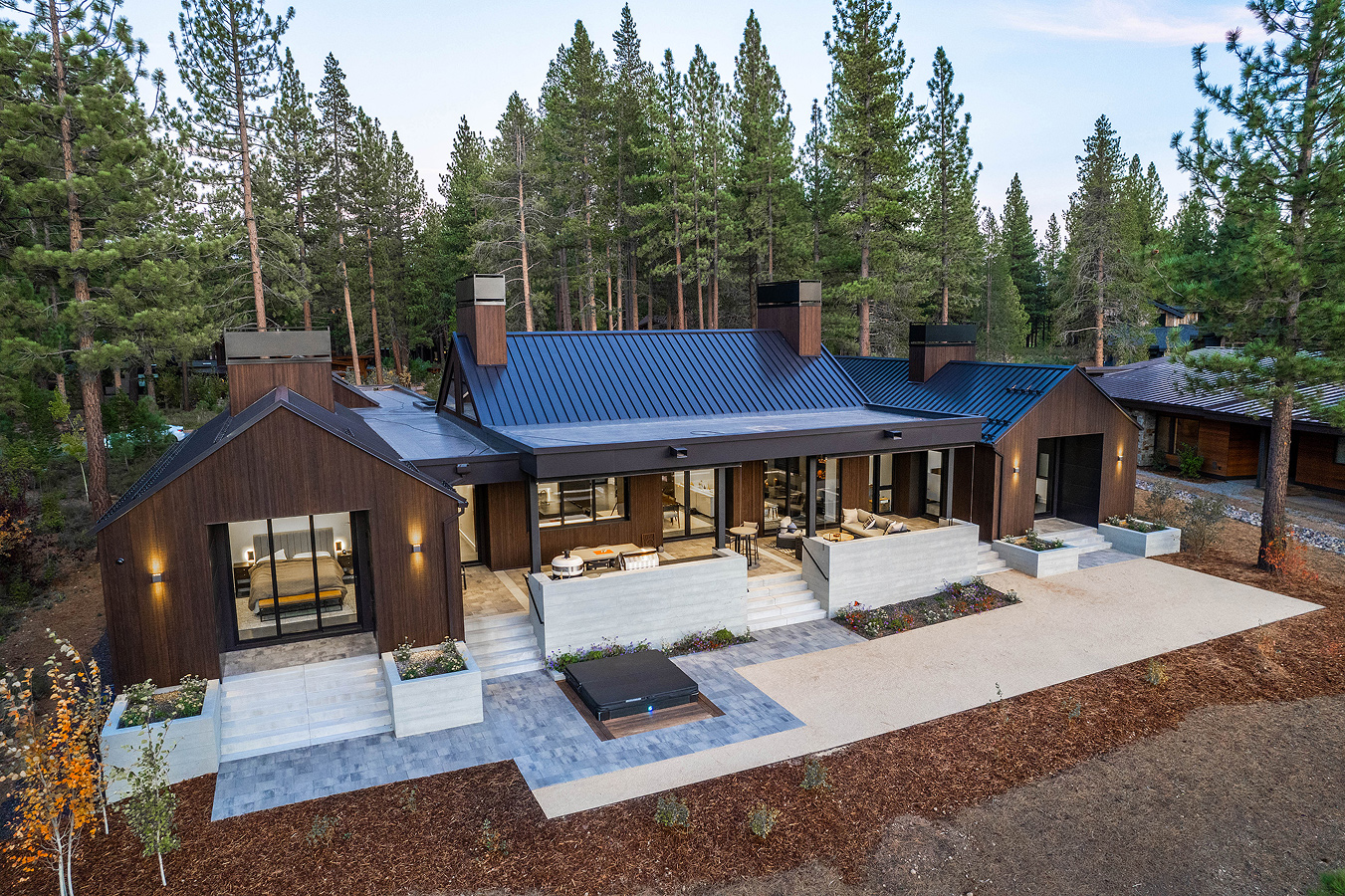 Modern single-story house with dark wood siding, black metal roof, outdoor patio areas, and surrounded by pine trees.