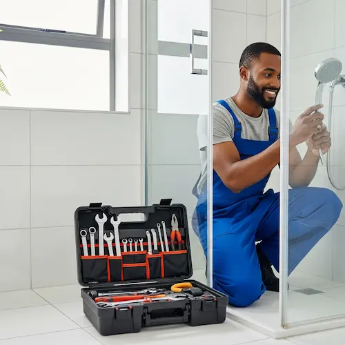 A smiling plumber in blue overalls kneels in a bathroom, fixing a showerhead.