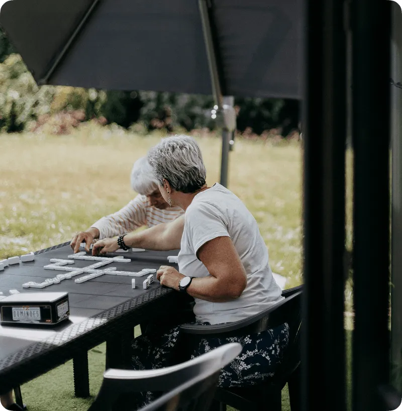 Deux personnes âgées jouant aux dominos assises à une table extérieure sous un parasol.
