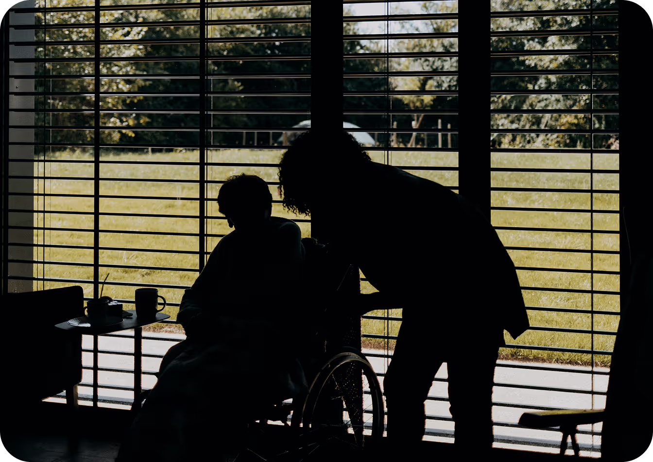 Silhouettes d'une personne en fauteuil roulant et d'une autre personne se penchant vers elle devant une fenêtre avec des stores, vue sur un jardin extérieur.