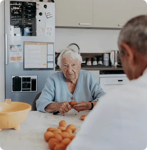 Une femme âgée coupant un abricot à une table de cuisine et un homme au premier plan flou.