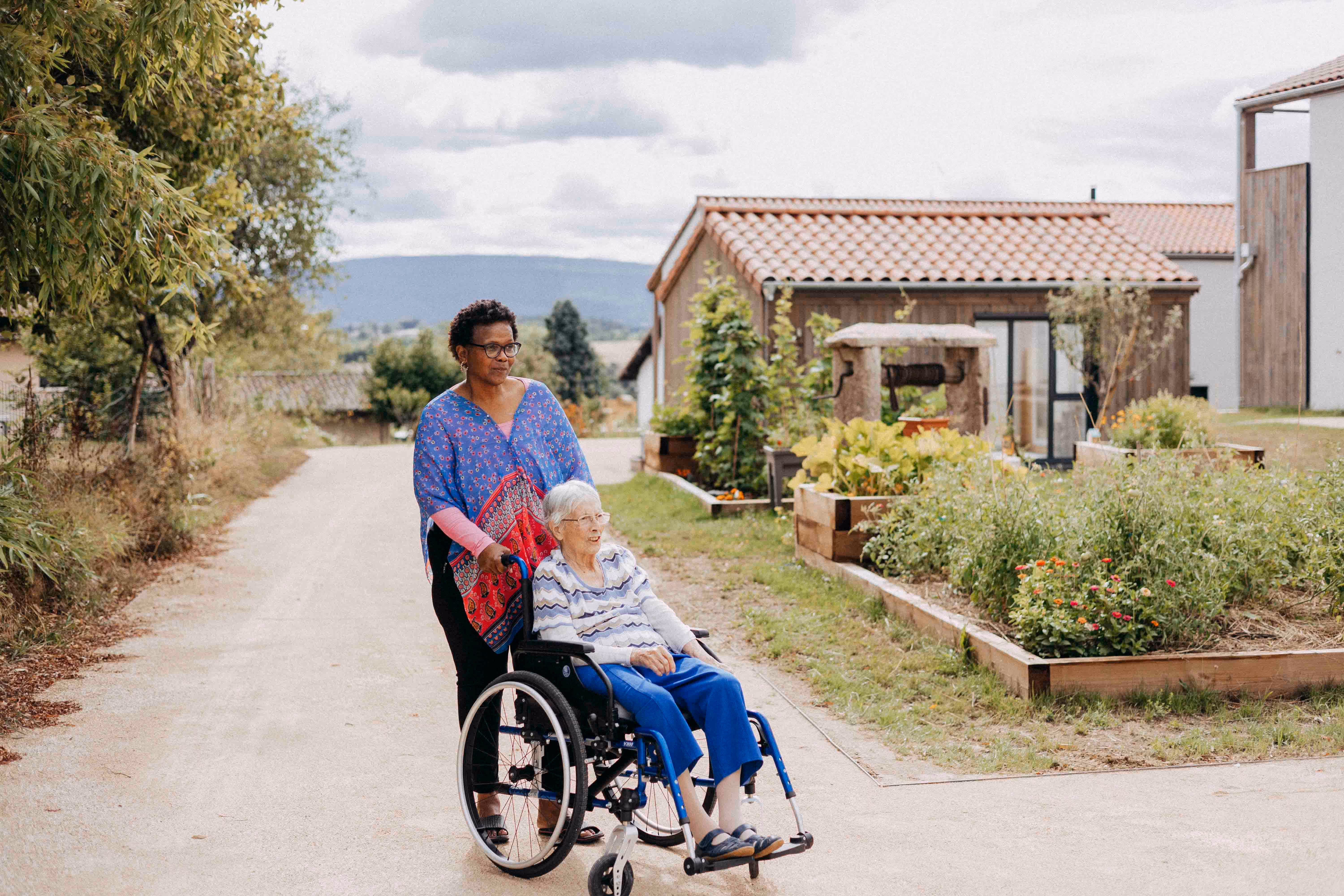 Une femme poussant une personne âgée en fauteuil roulant sur un chemin en plein air, avec des jardins surélevés et des maisons en arrière-plan.