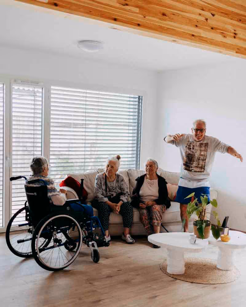 Quatre personnes âgées discutant dans un salon lumineux, trois assises dont une en fauteuil roulant et un homme debout faisant un geste joyeux.