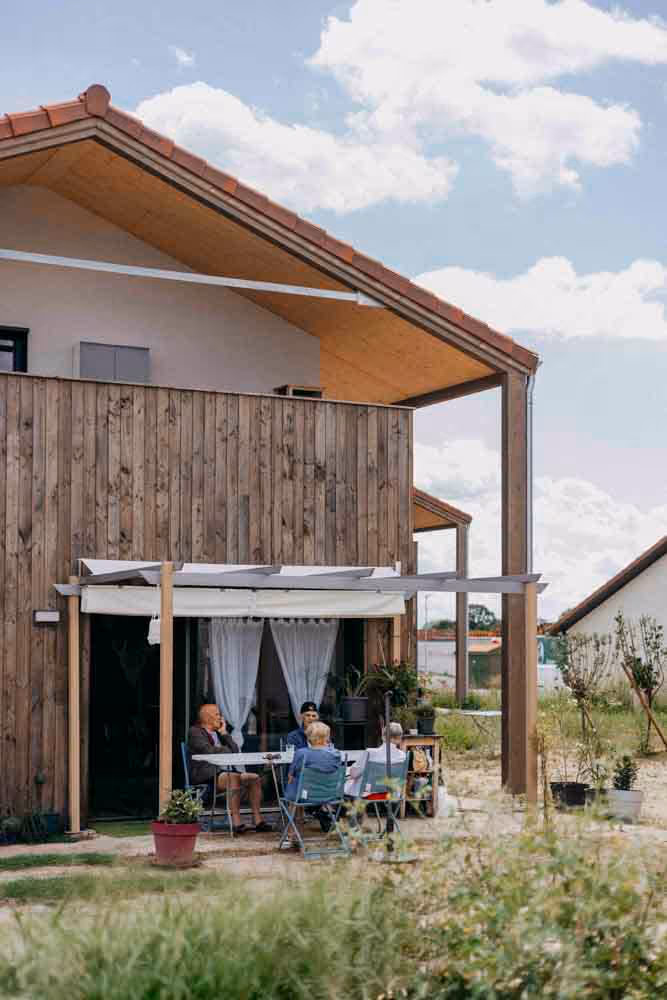 Quatre personnes âgées assises autour d'une table sous une pergola devant une maison en bois.