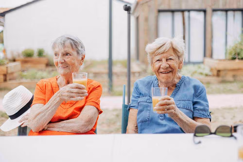 Deux femmes âgées souriantes assises à une table extérieure, tenant chacune un verre et trinquant