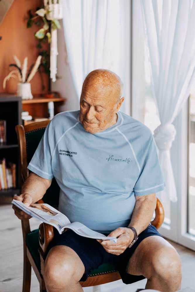 Homme âgé assis sur une chaise à la maison, lisant un livre près d'une fenêtre 