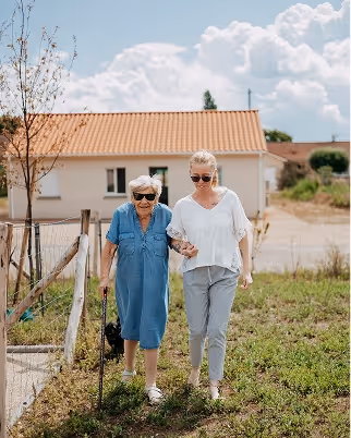 Une femme plus jeune aide une femme âgée avec une canne à marcher dans un jardin devant une maison. Elles sourient