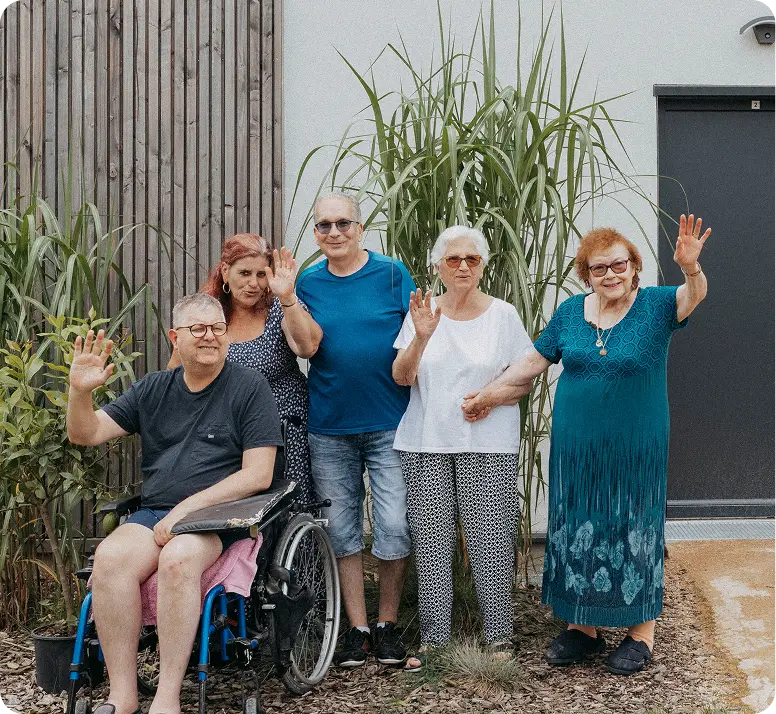 Un groupe de cinq personnes âgées, dont un homme en fauteuil roulant, sourient et lèvent la main devant un mur extérieur avec des plantes.