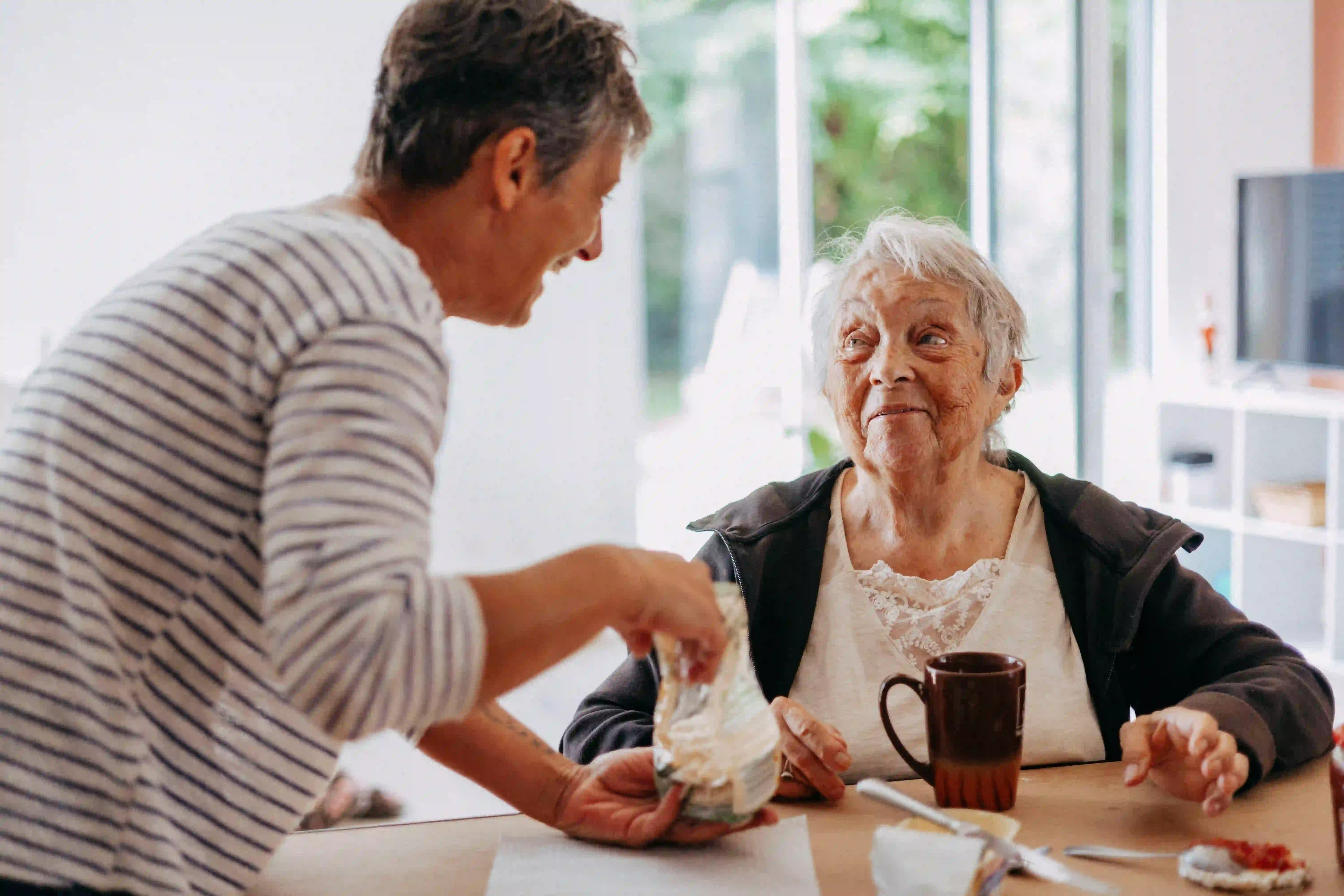 Une femme souriante tend un sac à une personne âgée assise à une table avec une tasse.