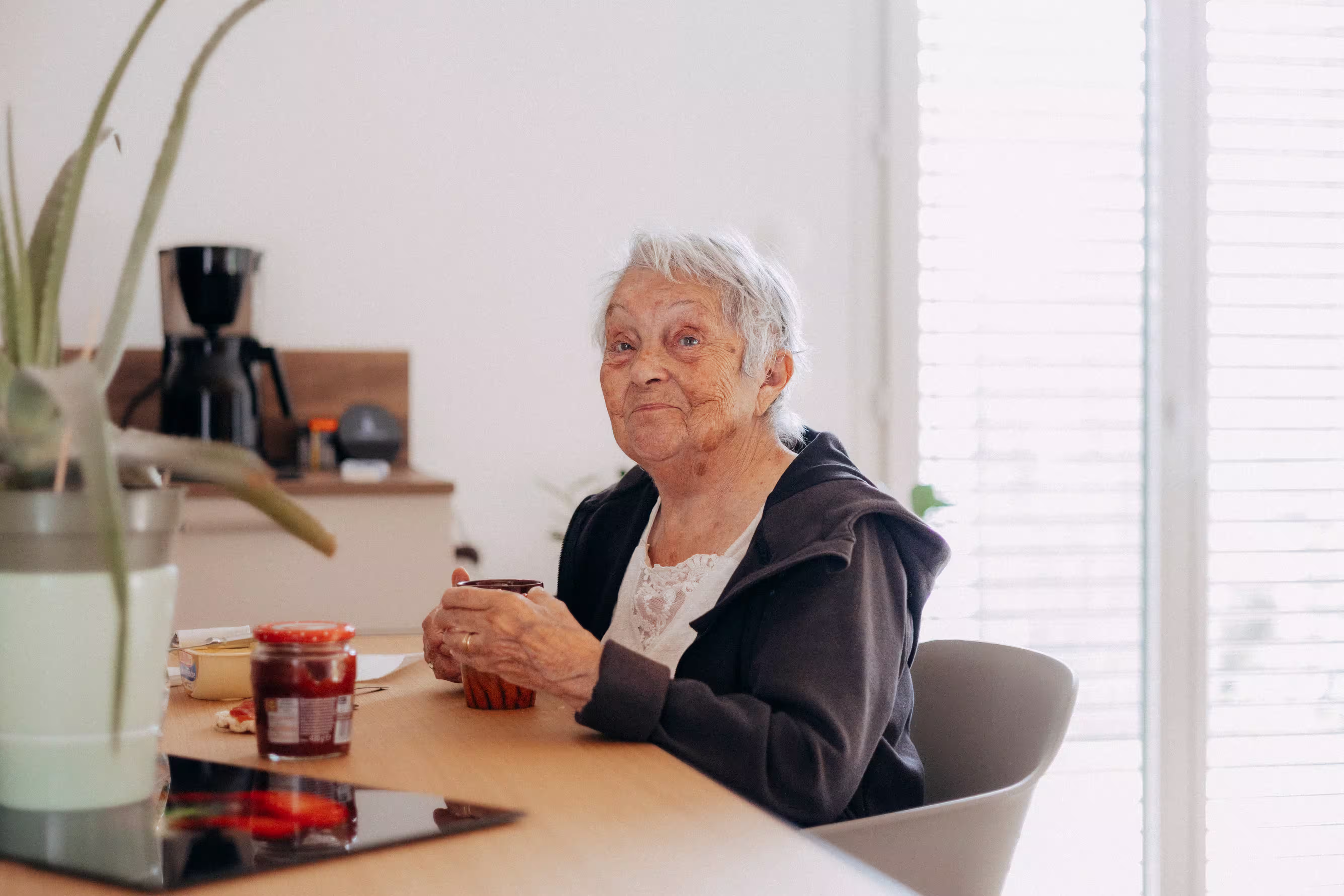 Femme âgée assise à une table avec une tasse, souriant dans une cuisine lumineuse.