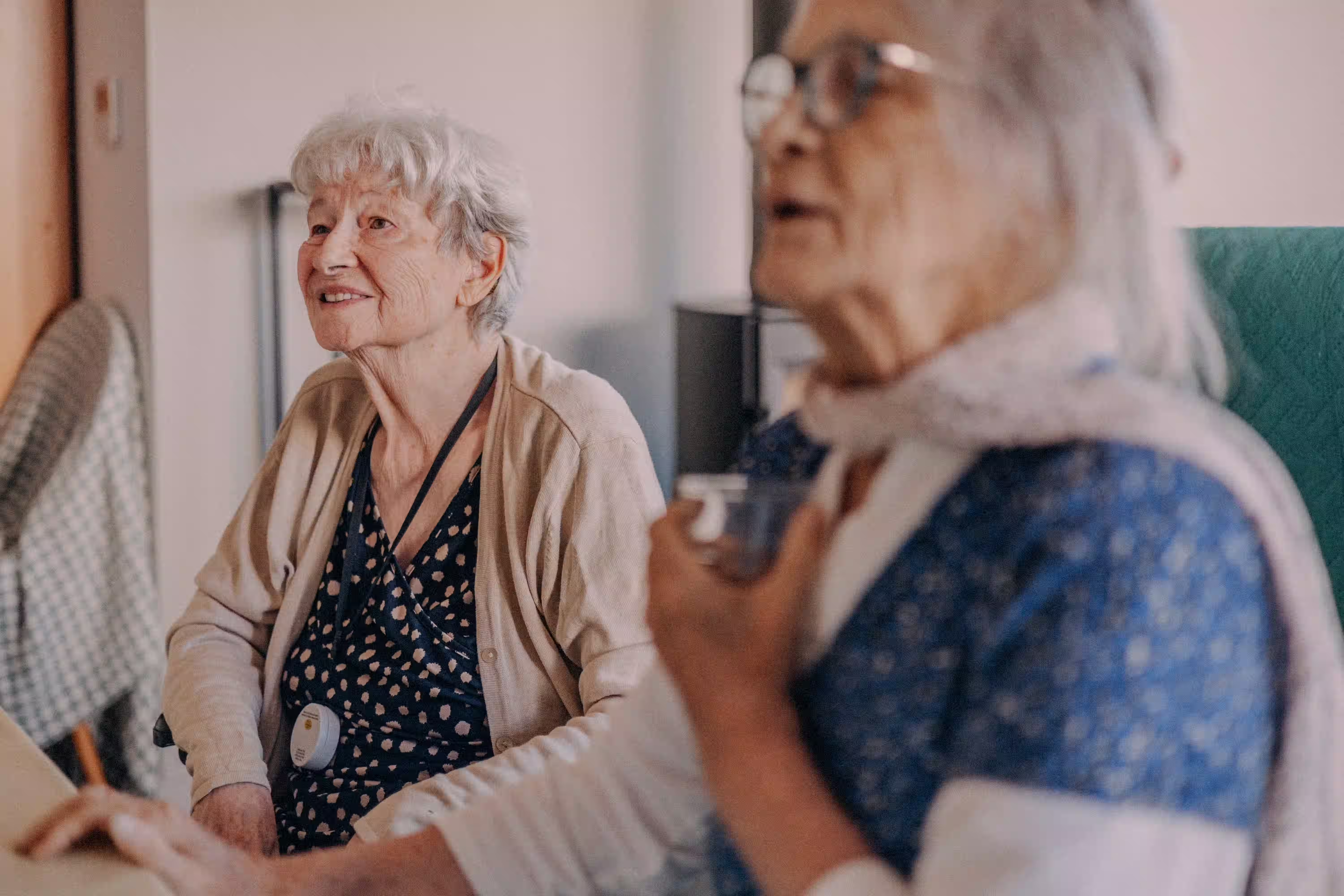 Deux femmes âgées assises, une souriante portant un gilet beige, l'autre floue tenant un verre.