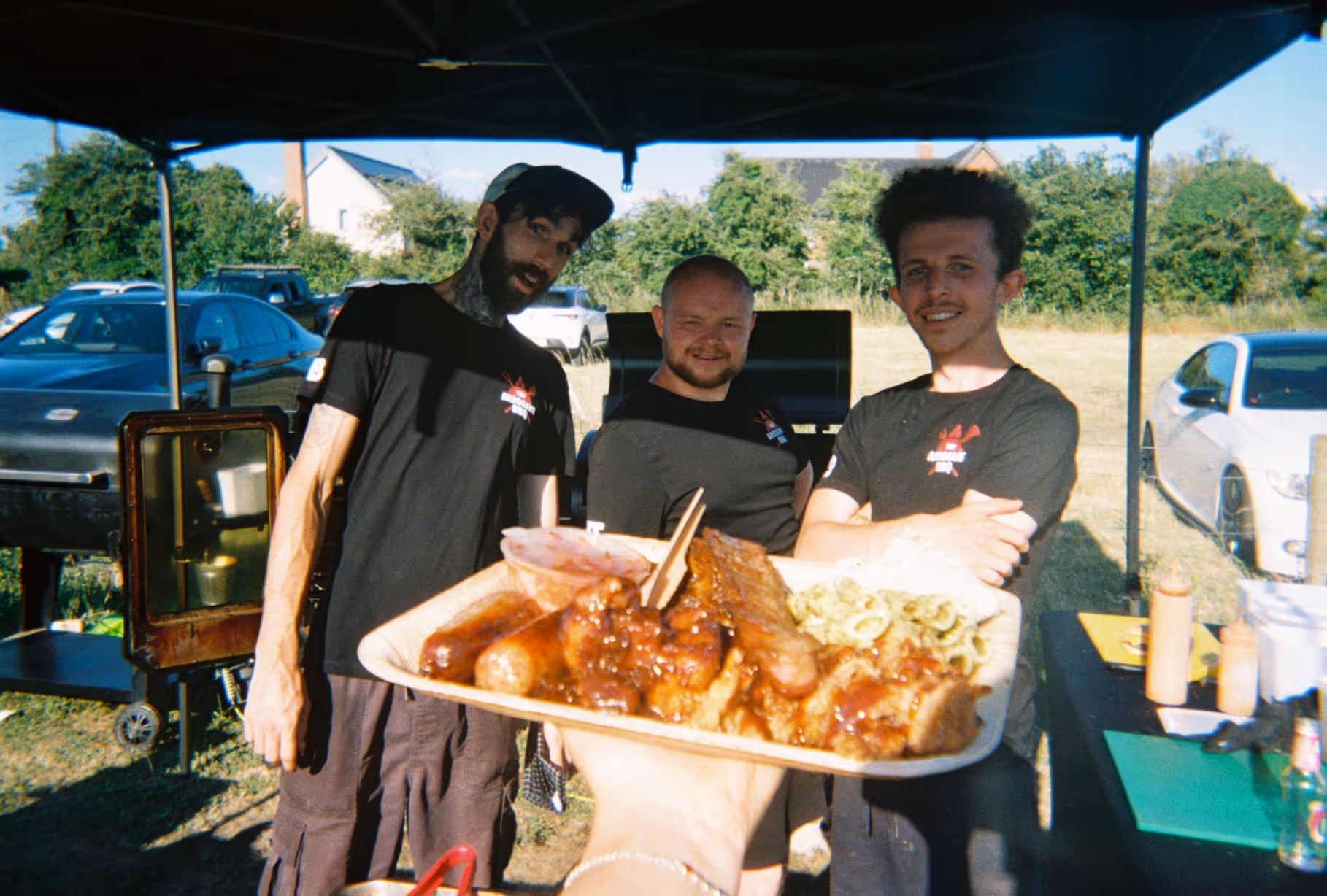 Three men in black shirts standing under a canopy outdoors, smiling behind a hand holding a plate of barbecued meats and sides.