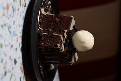 Square chocolate brownie topped with white chocolate drizzle, crumbled cookie pieces, and a scoop of vanilla ice cream on a black plate.