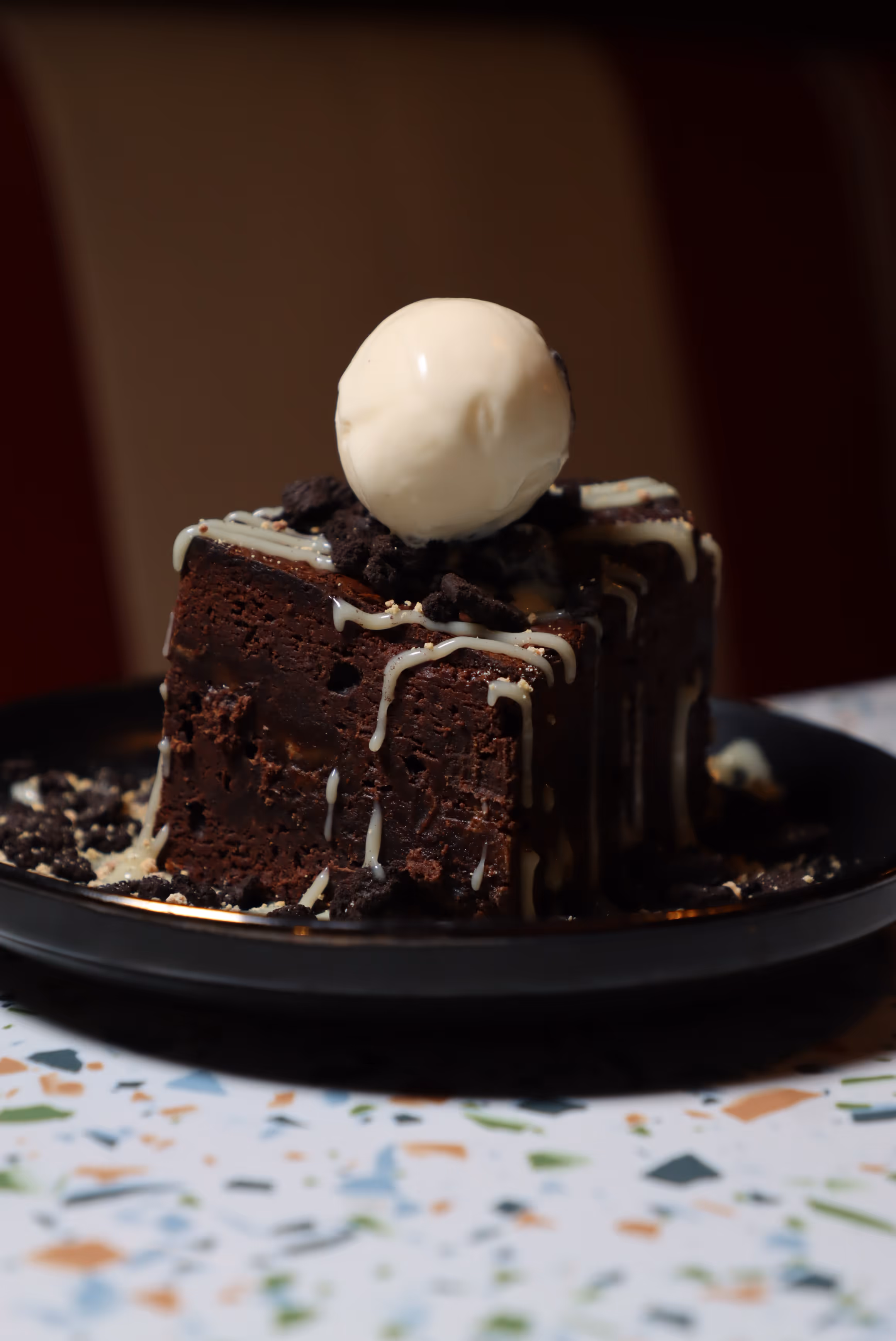 Square chocolate brownie topped with white chocolate drizzle, crumbled cookie pieces, and a scoop of vanilla ice cream on a black plate.