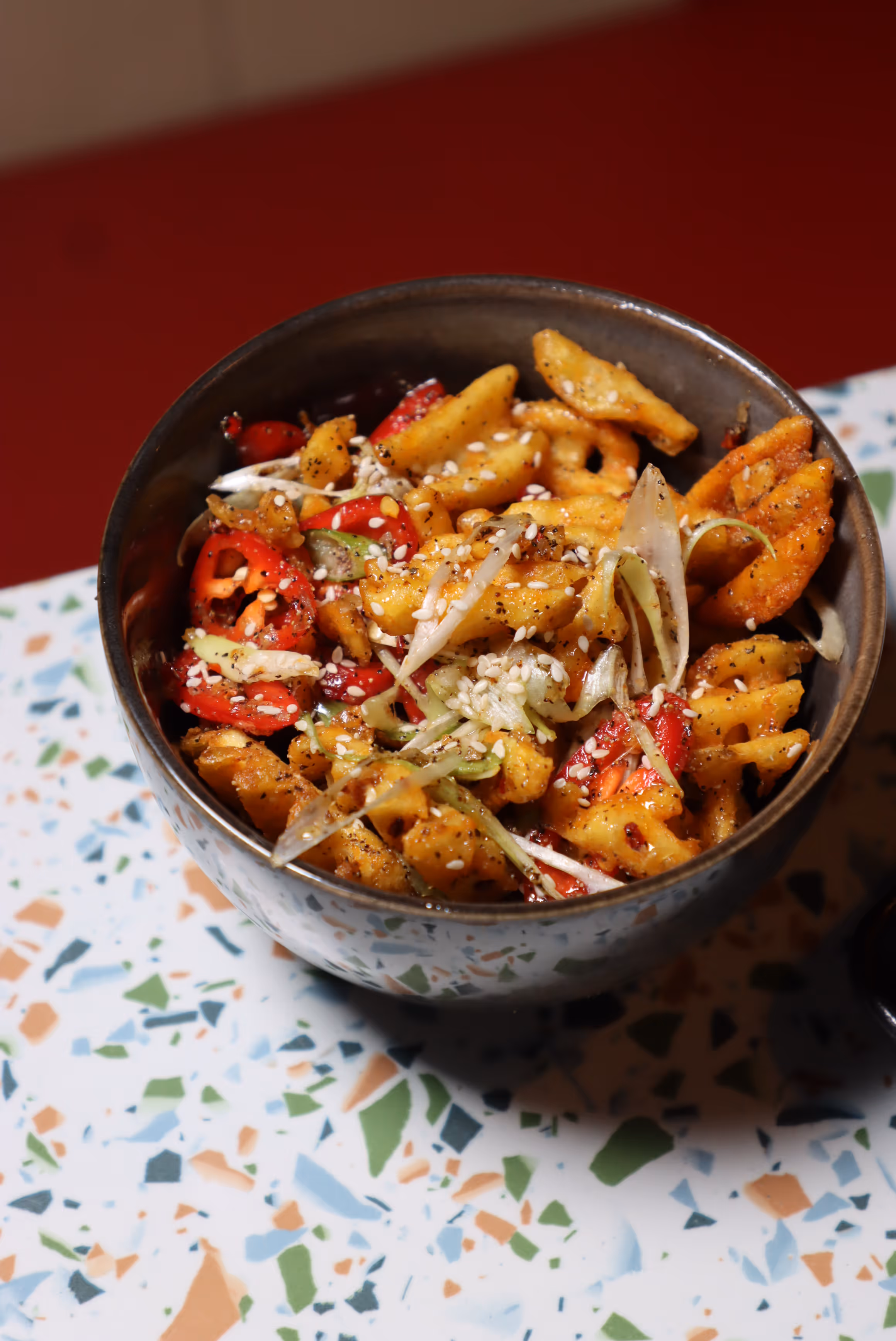 Bowl of seasoned waffle fries mixed with sliced red peppers, onions, and sprinkled sesame seeds on a terrazzo tabletop.