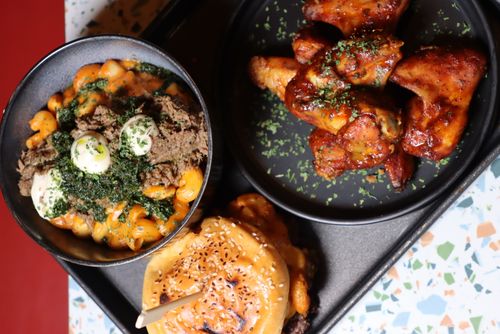 Tray with a bowl of creamy pasta topped with shredded meat, green sauce, and dollops of white sauce, a sesame-seeded bun sandwich, and a plate of seasoned chicken wings garnished with herbs.