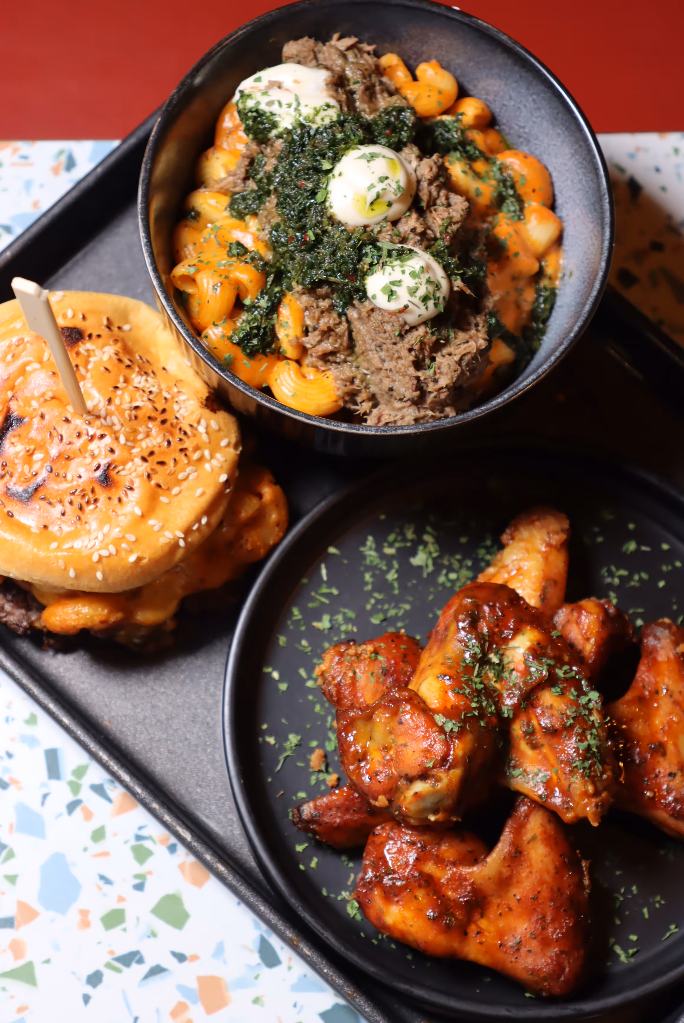 Tray with a bowl of creamy pasta topped with shredded meat, green sauce, and dollops of white sauce, a sesame-seeded bun sandwich, and a plate of seasoned chicken wings garnished with herbs.