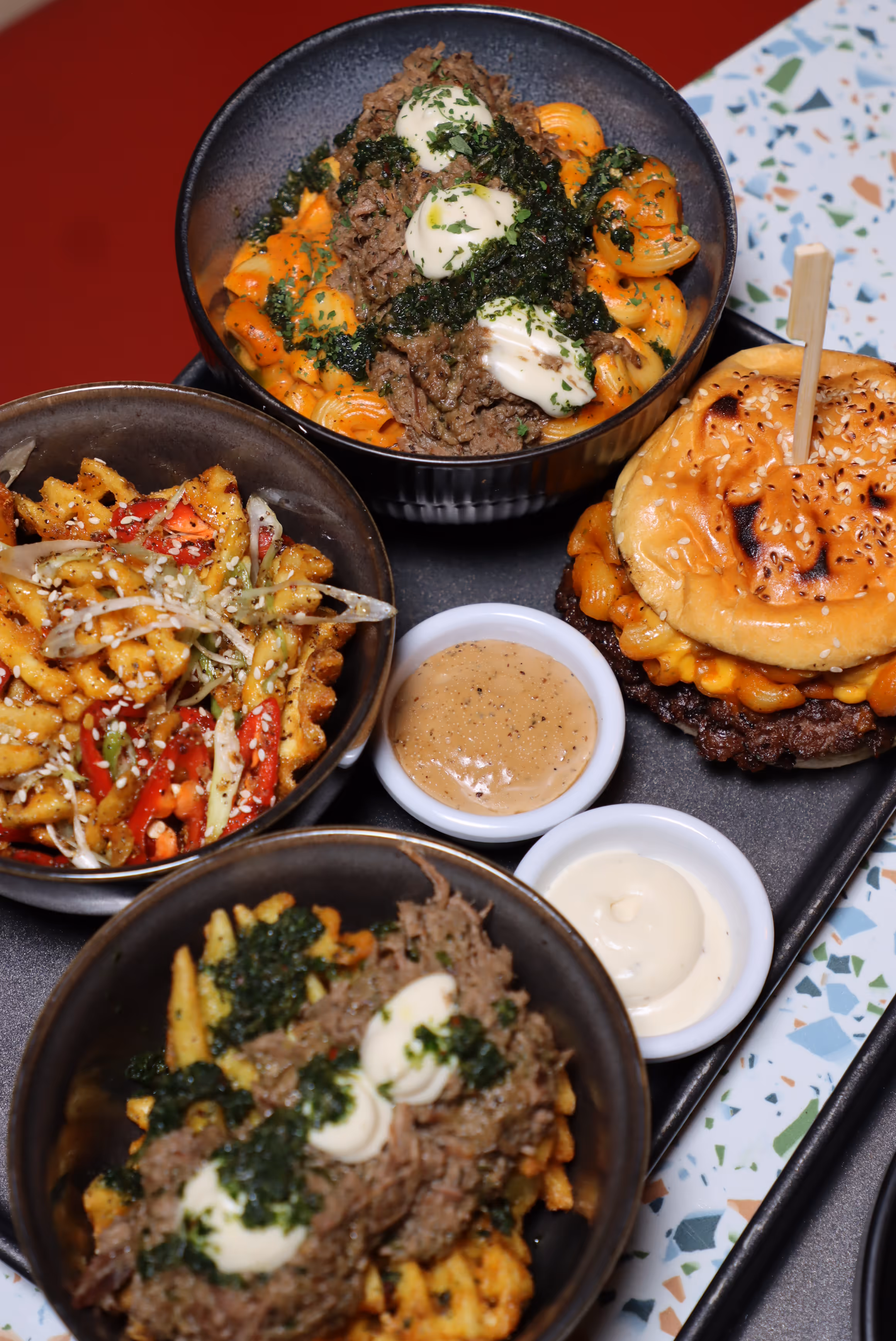 Tray with a cheeseburger topped with macaroni, two bowls of seasoned waffle fries with shredded meat and cream, a bowl of seasoned spiral pasta with shredded meat and cream, and two dipping sauces.