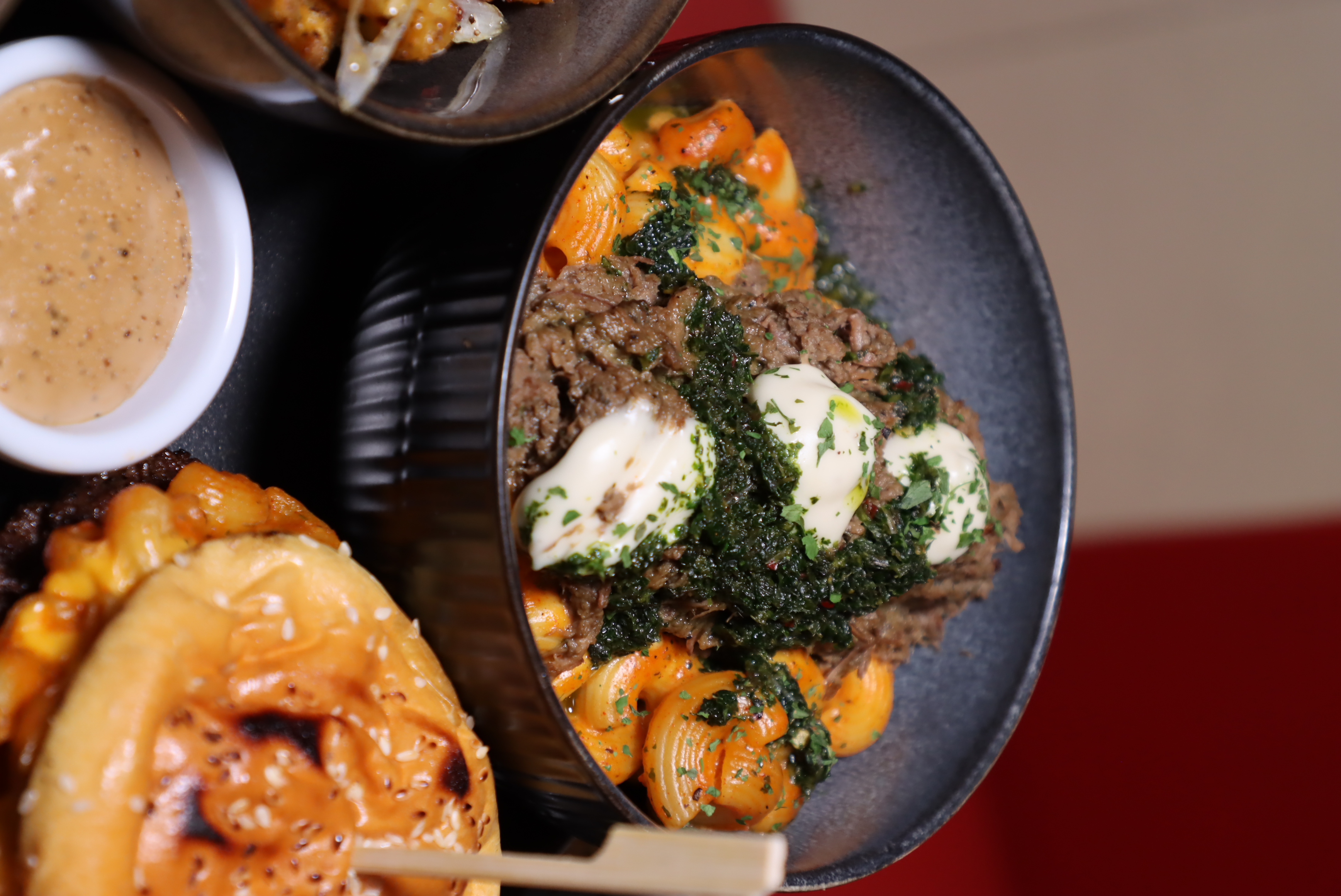 Bowl of shell pasta topped with shredded beef, creamy sauce dollops, and green herbs, next to a sesame seed bun sandwich and a small cup of dipping sauce.