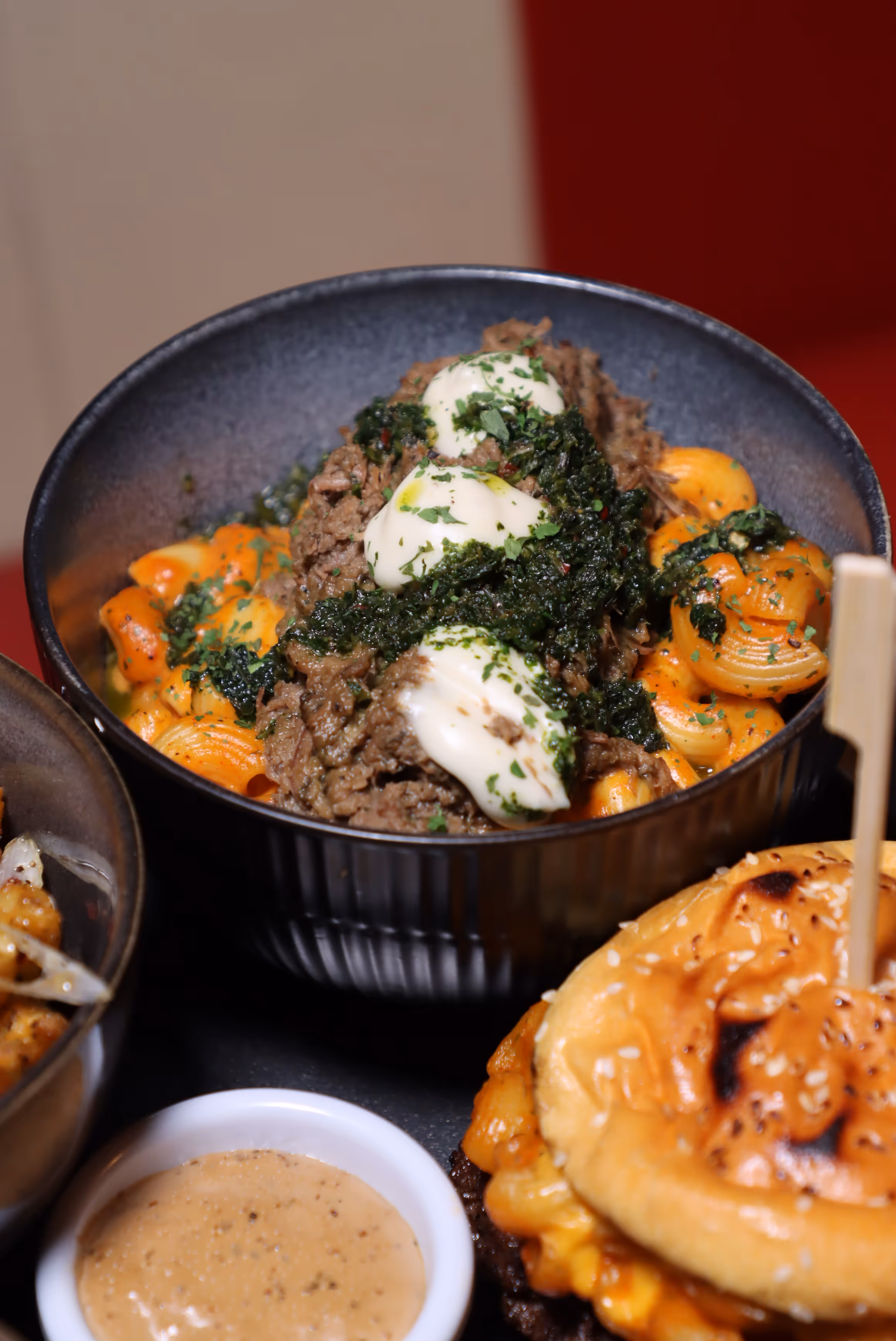 Bowl of shell pasta topped with shredded beef, creamy sauce dollops, and green herbs, next to a sesame seed bun sandwich and a small cup of dipping sauce.