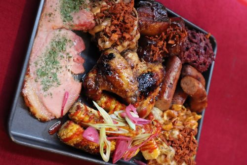 Tray with grilled chicken wings, sausages, ribs with sauce, corn on the cob with pink pickled onions and green garnish, sliced meat with herbs, and sides of macaroni and pulled meat topped with crispy bits.