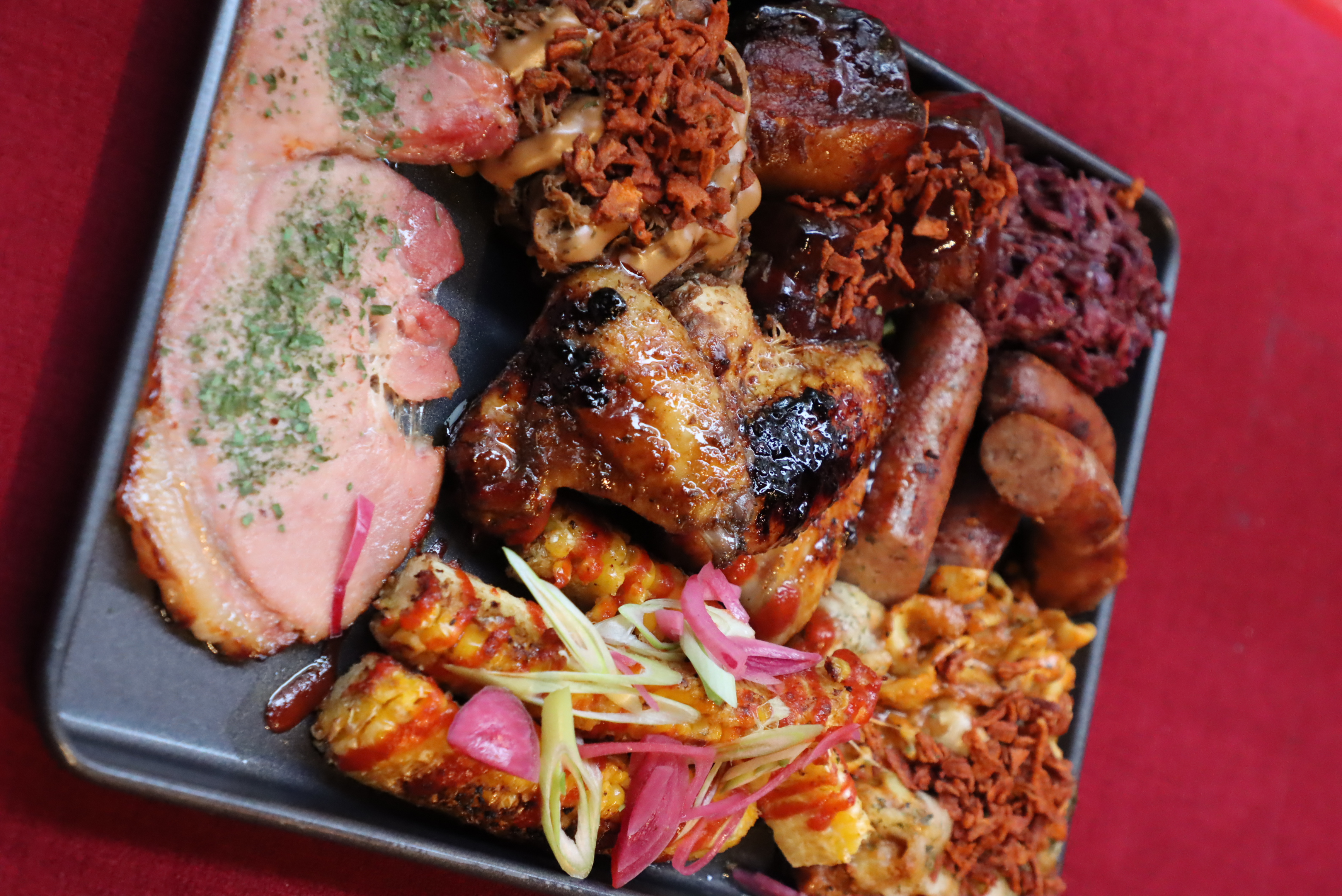 Tray with grilled chicken wings, sausages, ribs with sauce, corn on the cob with pink pickled onions and green garnish, sliced meat with herbs, and sides of macaroni and pulled meat topped with crispy bits.