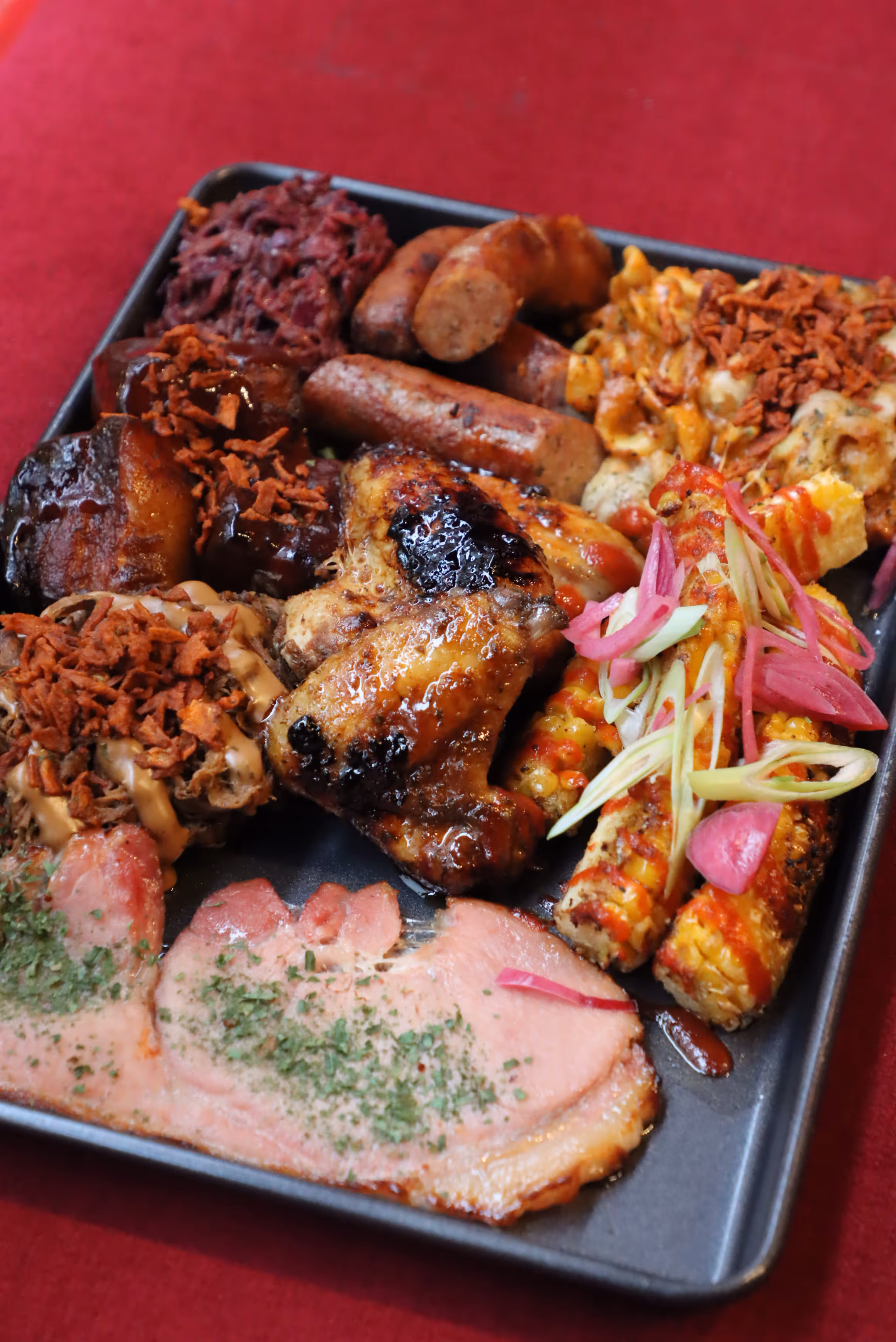 Tray with grilled chicken wings, sausages, ribs with sauce, corn on the cob with pink pickled onions and green garnish, sliced meat with herbs, and sides of macaroni and pulled meat topped with crispy bits.