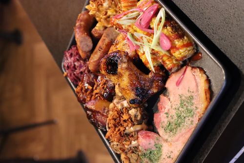 Plate with grilled chicken wing, sausages, corn on the cob with toppings, sliced ham, and other barbecued meats with crispy fried onions.