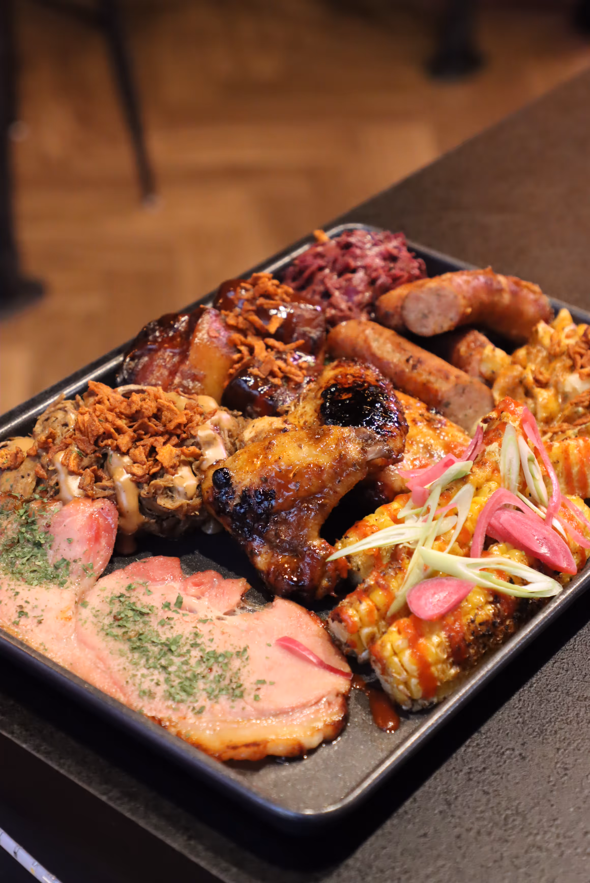 Plate with grilled chicken wing, sausages, corn on the cob with toppings, sliced ham, and other barbecued meats with crispy fried onions.