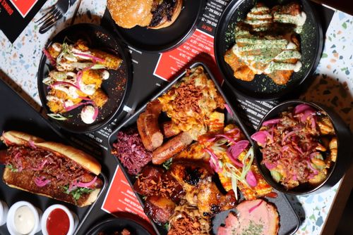 Variety of grilled and sauced dishes including a loaded hot dog, corn on the cob with toppings, a burger, chicken wings with sauce, sausages, and poutine with pickled onions on black plates and trays.